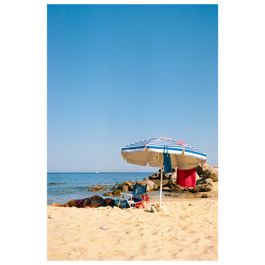 A person lying under a parasol at the beach. They are in the shade, while the rest of the scene is bathed in a bright warm light. Various items are hanging from the parasol.