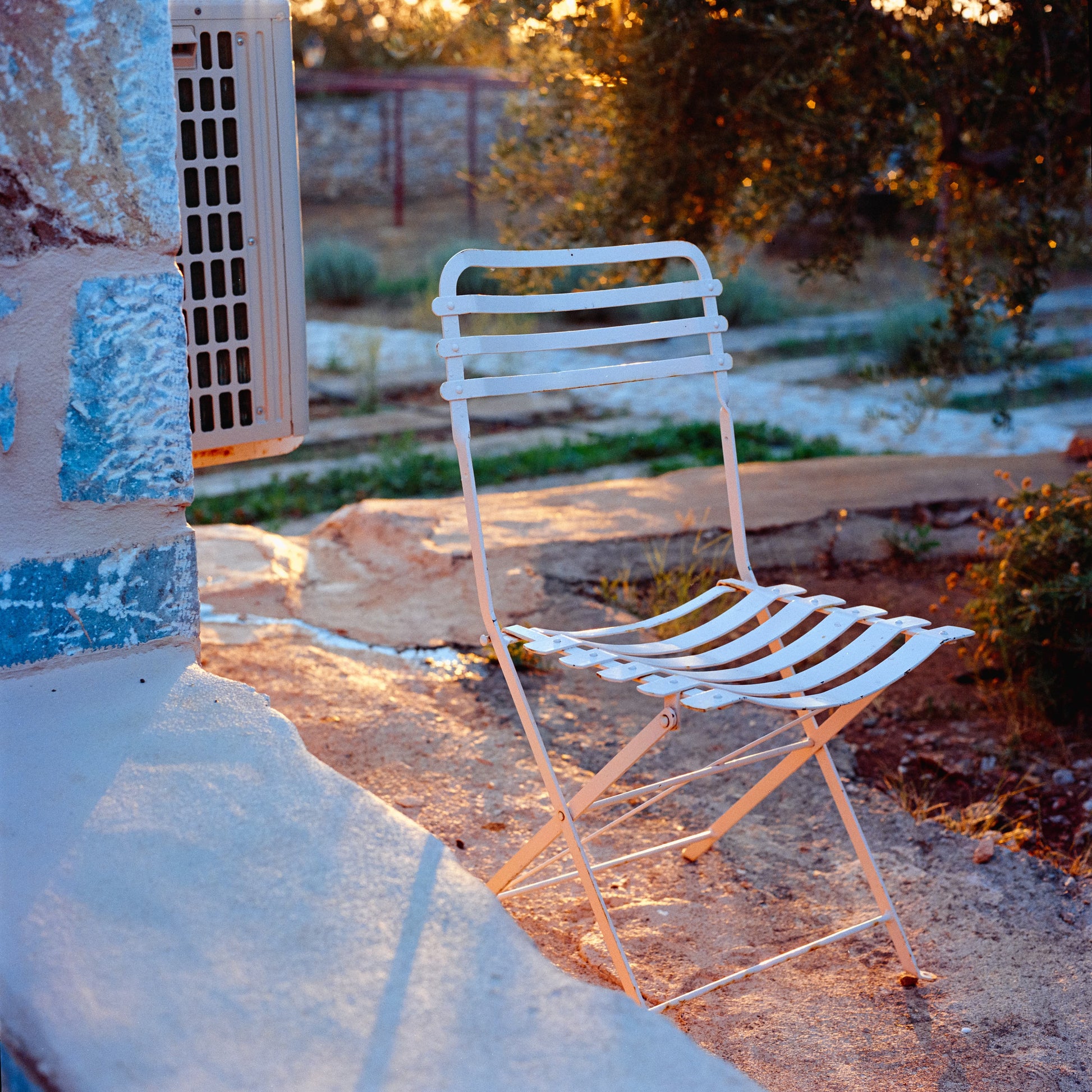 A square frame with a white metal chair with grills standing in the middle. Itss eat and back trace a similar shape as a stone porch on the foreground. The sun casts rays down the middle of the frame, and small shadows from the chair. There is a warmth and stillness in the scene.