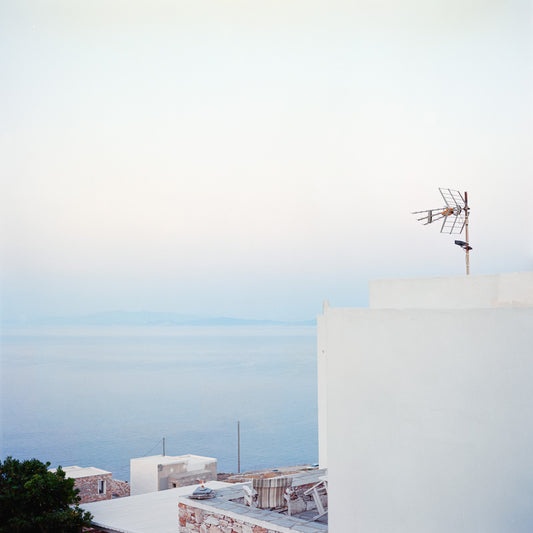 A square frame over white buildings and the sea. The dominant colourse are a light blue and white. On a building on the right, a TV antenna is pointing towards the distance.
